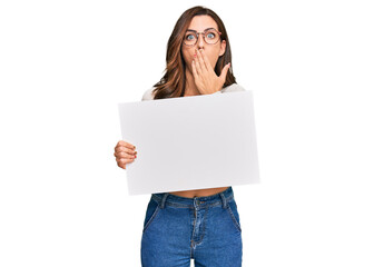 Young brunette woman holding blank empty banner covering mouth with hand, shocked and afraid for mistake. surprised expression