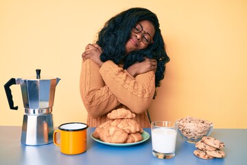 Beautiful african woman sitting on the table eating breakfast in the morning hugging oneself happy...