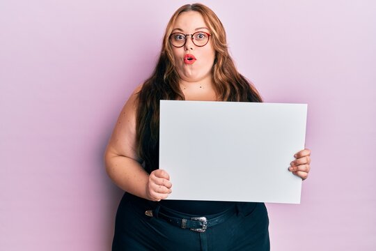 Plus size caucasian young woman holding blank empty banner scared and amazed with open mouth for surprise, disbelief face