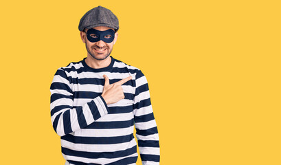 Young handsome man wearing burglar mask cheerful with a smile on face pointing with hand and finger up to the side with happy and natural expression