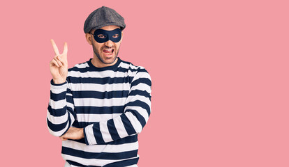 Young handsome man wearing burglar mask smiling with happy face winking at the camera doing victory sign with fingers. number two.