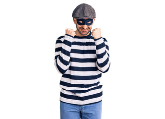 Young handsome man wearing burglar mask excited for success with arms raised and eyes closed celebrating victory smiling. winner concept.