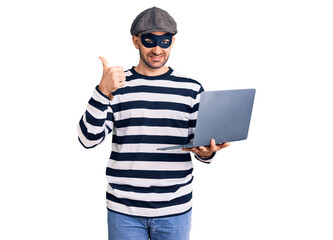 Young handsome man wearing burglar mask using laptop smiling happy and positive, thumb up doing excellent and approval sign