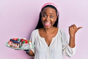 Young african woman holding plate with cake slices pointing thumb up to the side smiling happy with open mouth