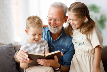 Cheerful grandfather and grandchildren reading book. - obrazy, fototapety, plakaty