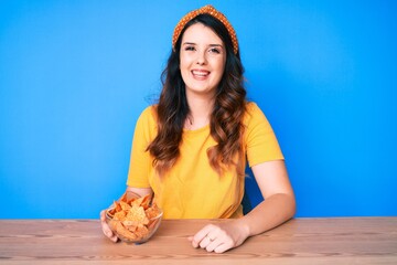 Young beautiful brunette woman sitting on the table eating nachos potato chips looking positive and...