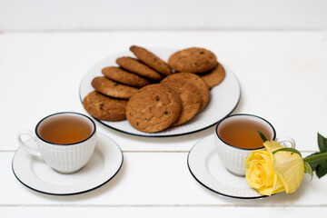 Two cups of tea and oatmeal cookies. Yellow rose. Breakfast. Tea drinking. White wooden background.