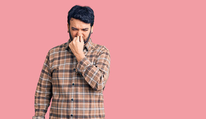 Young hispanic man wearing casual clothes smelling something stinky and disgusting, intolerable smell, holding breath with fingers on nose. bad smell