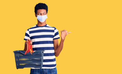 Young african american man wearing shopping basket and medical mask pointing thumb up to the side smiling happy with open mouth