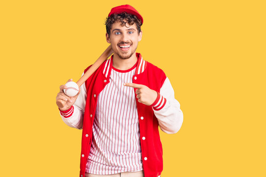 Young handsome man with curly hair playing baseball holding bat and ball smiling happy pointing with hand and finger