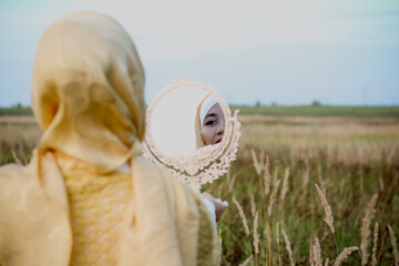 A Muslim woman stands on the field and holds a mirror in her hands. Muslim woman in beige hijab. Beauty, femininity, feminism