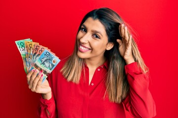 Beautiful brunette woman holding australian dollars smiling with hand over ear listening an hearing to rumor or gossip. deafness concept.