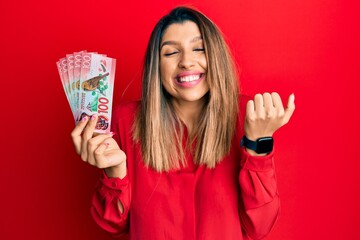 Beautiful brunette woman holding 100 new zealand dollars banknote screaming proud, celebrating victory and success very excited with raised arm