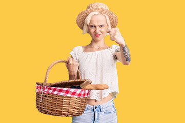 Young blonde woman with tattoo wearing summer hat and holding picnic wicker basket with bread annoyed and frustrated shouting with anger, yelling crazy with anger and hand raised