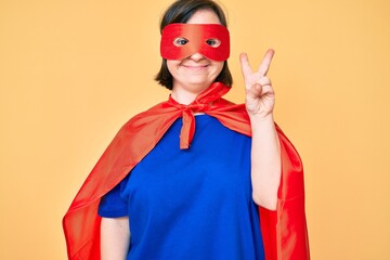 Brunette woman with down syndrome wearing super hero costume smiling with happy face winking at the camera doing victory sign. number two.