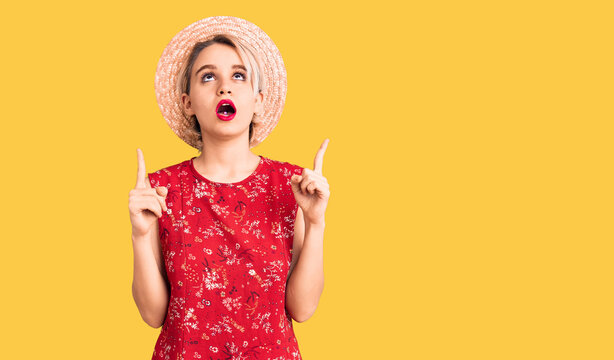 Young beautiful blonde woman wearing summer hat amazed and surprised looking up and pointing with fingers and raised arms.