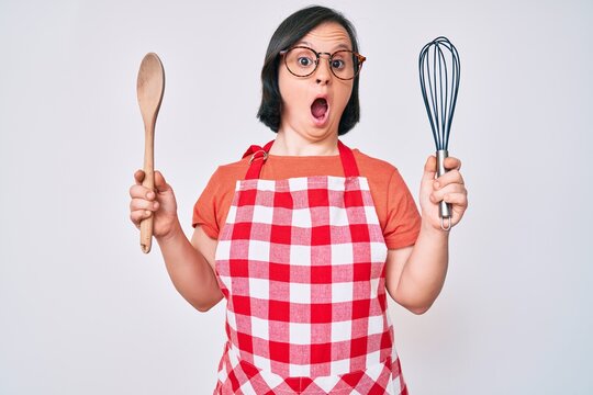 Brunette woman with down syndrome cooking using baker whisk and spoon afraid and shocked with surprise and amazed expression, fear and excited face.