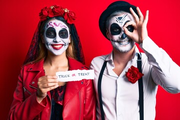 Couple wearing day of the dead costume holding trick or treat paper smiling happy doing ok sign with hand on eye looking through fingers