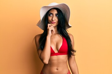 Beautiful hispanic woman wearing bikini and summer hat thinking concentrated about doubt with finger on chin and looking up wondering