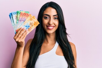 Beautiful hispanic woman holding swiss franc banknotes looking positive and happy standing and smiling with a confident smile showing teeth