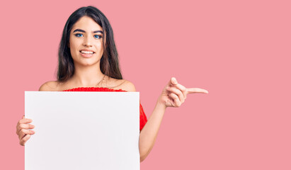 Brunette teenager girl holding blank empty banner smiling happy pointing with hand and finger to the side