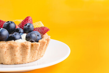 Acercamiento tartaleta tropical de fresas, melocotón y arándanos frescos sobre un plato de cerámica blanco sobre fondo amarillo ideal para redes sociales o publicidad.