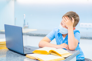 A boy studies remotely near a laptop in a medical mask, leaning over open books at home in the...