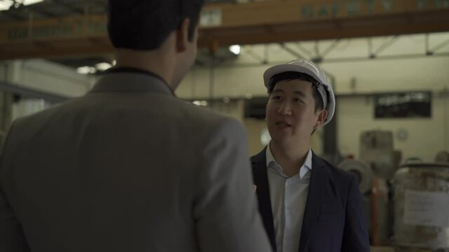30s young Asian businessman in formal suit and hard hat shaking hands with Indian business partner in factory background. Business partnership and team work success concept - 4K Real Time Footage