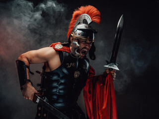 Brutal and savage roman soldier in dark armour with helmet and red cloak screams and assaults holding dual swords in smokey background.