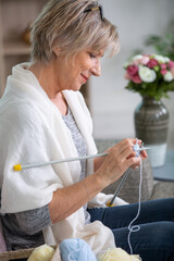 senior woman knitting warm scarf in armchair at home