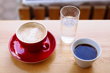 Aromatic coffee in a red cup with milk foam and latte art and freshly brewed coffee in a white cup. Nearby is a glass of water. Enjoyment of hot aroma beverage