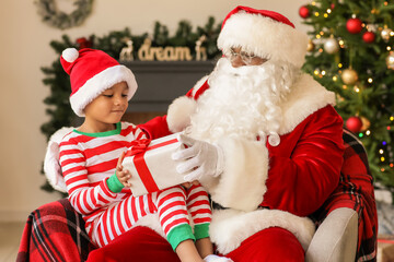 Fototapeta na wymiar African-American Santa Claus and cute boy with gift at home on Christmas eve