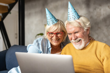 Cheerful modern caucasian elderly couple making video call on laptop for birthday or anniversary celebration while sitting on sofa at home. Videoconference holiday greetings concept