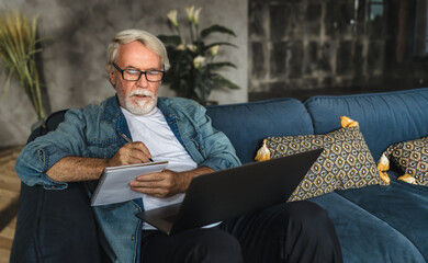 Focused elderly man writing notes in notebook watching webinar or online training using laptop...