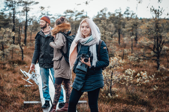 Attractive blond girl with camera poses at wooden path looking at camera behind her friends in lovely and relaxed autumn forest.