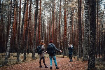 Fototapeta premium Blond girl with camera photographing her friends they stay together on path around trees in autumn forest.