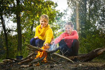 mother and daughter in the autumn forest sitting by the fire. mom and daughter making fire in woods.