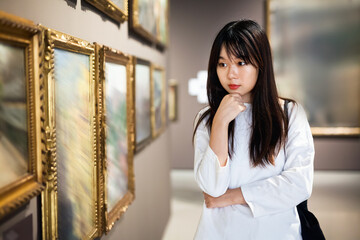 Serious positive glad chinese female visitor looking at artwork painting in the museum indoors