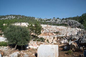 Fototapeta premium Marble industrial quarry, cutting of white and gray marble in natural environment with rocks, trees and blue sky and leaving their wastes to nature