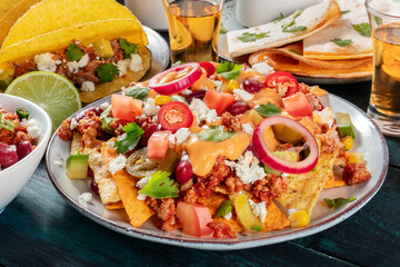 Mexican food. Nachos with beef, beans, chili peppers, onions and cheese sauce close-up, with pork tacos, quesadillas and tequila in the background, on a dark wooden table
