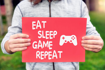 Male teenager gamer holds a banner with eat, sleep, game, repeat quote message with a gamepad symbol.