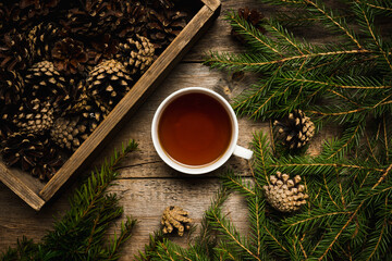 Christmas hygge background with cup of tea, pine branches and crate of cones on the wooden...