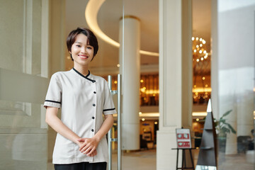 Portrait of cheerful young Asian hotel manager standing at entrance doors and smiling at camera