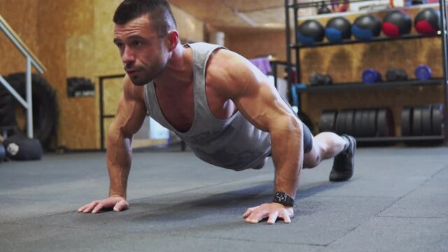 Concentrated muscular man doing push-ups on floor at the  gym, dressed in gray sportswear. Fitness club body weight workout. Wide shot.