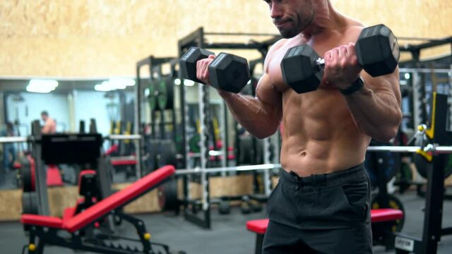 Fitness man with naked torso pumping up arm muscles. Bodybuilding workout in a gym. Muscular athlete doing biceps curls exercises with heavy dumbbells. Handsome power guy is training hard.
