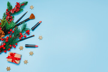 red lipsticks, makeup brushes and a gift lie on a blue background next to spruce branches with red berries. cosmetics for festive makeup for Christmas. Winter sales of cosmetics. Copy space, top view