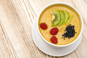 Photo of mango smoothie with raspberries, kiwi slices and chia seeds in a white bowl on a wooden background. Healthy organic food.