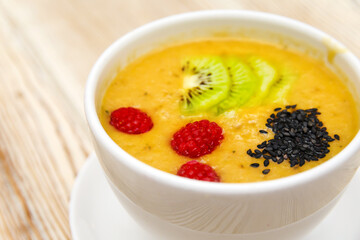 Closeup photot of mango smoothie with kiwi slices, raspberries and chia seeds in a white bowl on a wooden background. Healthy organic food. Selective focus