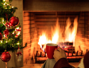 Hand holding a mug of tea, logs burning in a fireplace