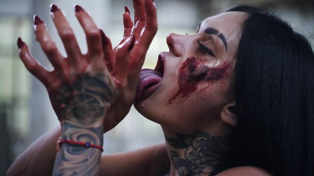 Zombie woman with bloody wound on her face licks her hand with tongue